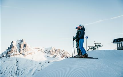 Two skiers are standing on a snow-covered slope with a view of impressive mountains. The sky is clear and sunny.