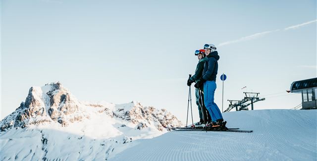 Zwei Skifahrer stehen auf einer schneebedeckten Piste mit Blick auf beeindruckende Berge. Der Himmel ist klar und sonnig.