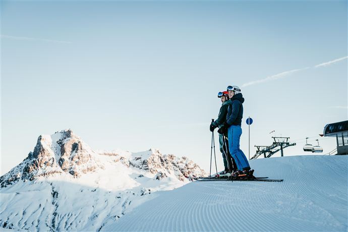 Zwei Skifahrer stehen auf einem verschneiten Hang und genießen die Aussicht auf die Berge. Der Himmel ist klar und blau.