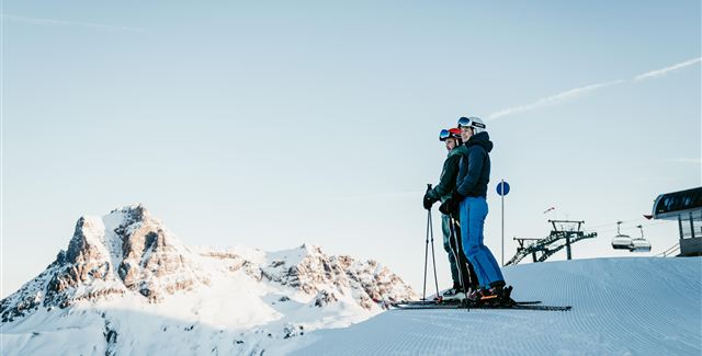 Two skiers are standing on a snowy slope and enjoying the view of the mountains. The sky is clear and blue.