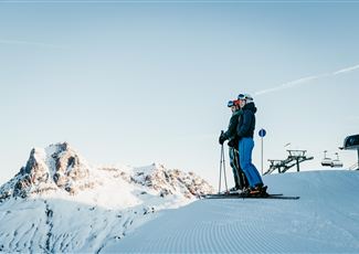 Two skiers are standing on a snowy slope and enjoying the view of the mountains. The sky is clear and blue.
