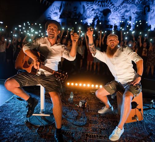 A live band is playing on a stage in the mountains. In the background, there are snow-covered peaks and spectators enjoying the concert.