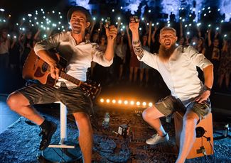 A live band is playing on a stage in the mountains. In the background, there are snow-covered peaks and spectators enjoying the concert.