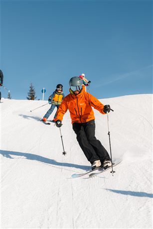 A group of skiers preparing on a snow-covered slope. The sun is shining and the mountains are visible in the background.