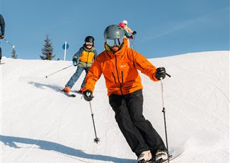 A group of skiers preparing on a snow-covered slope. The sun is shining and the mountains are visible in the background.