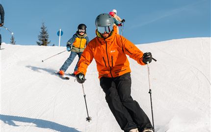 Eine Gruppe von Skifahrern, die sich auf einem schneebedeckten Hang vorbereitet. Die Sonne scheint und die Berge sind im Hintergrund sichtbar.