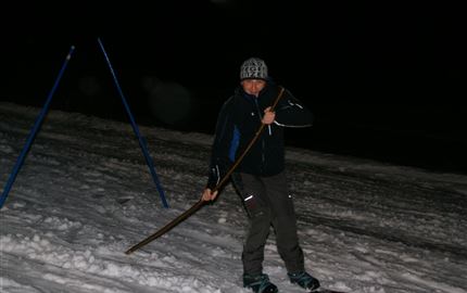 Ein Skifahrer bei Nacht auf einer schneebedeckten Piste. Er hält einen Stock in der Hand und trägt passende Ski-Ausrüstung.