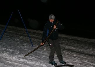 A skier at night on a snow-covered slope. He is holding a pole in his hand and wearing appropriate skiing gear.
