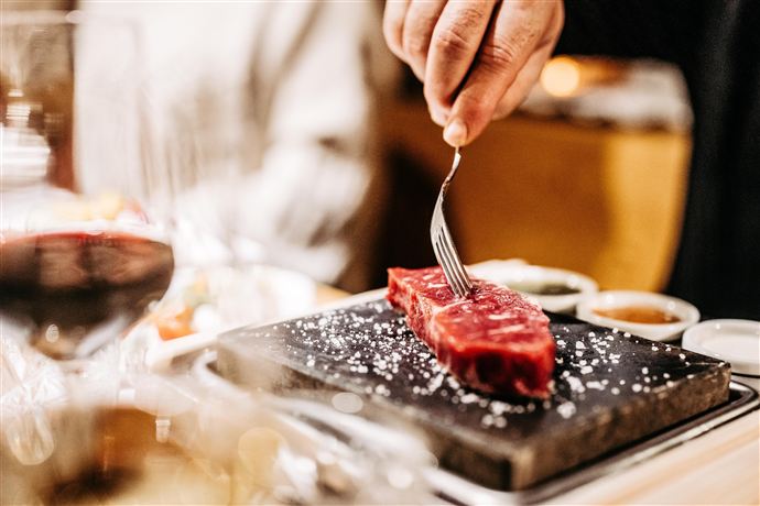 Ein Stück rotes Fleisch wird mit einer Gabel auf einem heißen Stein zubereitet. Im Hintergrund sind Gläser und Saucen zu sehen.