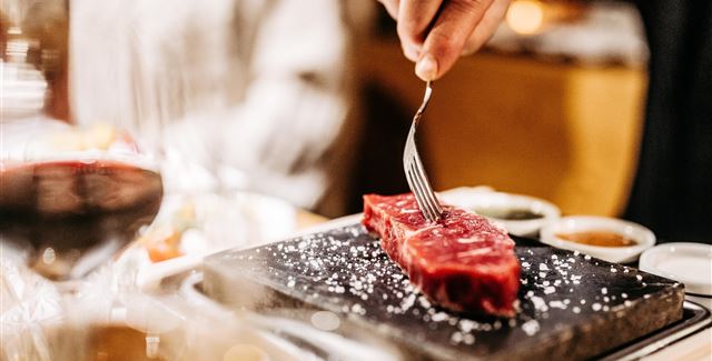 Ein Stück rotes Fleisch wird mit einer Gabel auf einem heißen Stein zubereitet. Im Hintergrund sind Gläser und Saucen zu sehen.