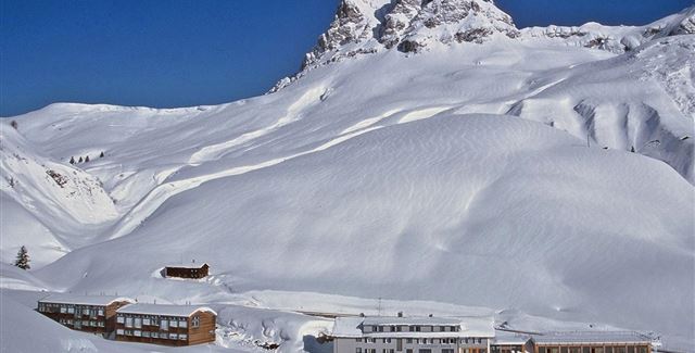 Eine schneebedeckte Landschaft mit einem gemütlichen Hotel und majestätischen Bergen im Hintergrund. Der Himmel ist klar und blau, was eine ruhige Winteratmosphäre schafft.