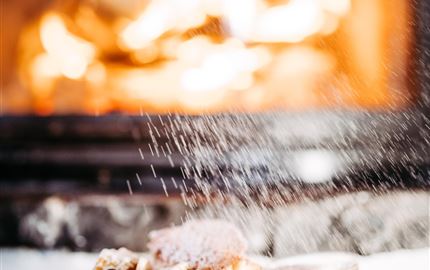 Ein Teller mit köstlichen, mit Puderzucker bestäubten Leckereien steht vor einem prasselnden Kamin. Der warme, gemütliche Hintergrund sorgt für eine einladende Atmosphäre.