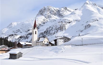 A snowy landscape with a small village and a church. In the background, majestic mountains rise.