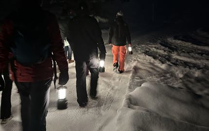 Eine Gruppe von Menschen wandert nachts mit Laternen durch eine schneebedeckte Landschaft. Die Umgebung ist dunkel und es ist ruhig.