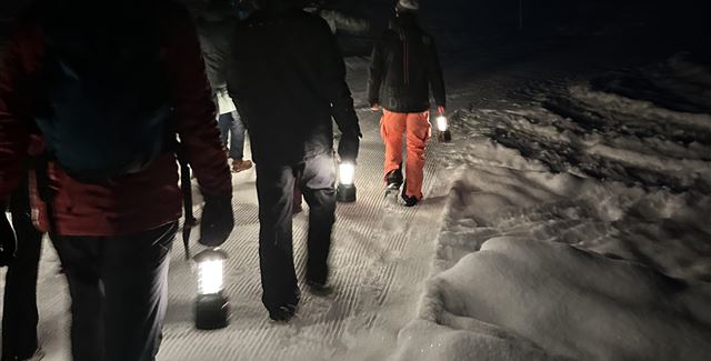 Eine Gruppe von Menschen wandert nachts mit Laternen durch eine schneebedeckte Landschaft. Die Umgebung ist dunkel und es ist ruhig.