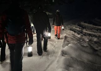 Eine Gruppe von Menschen wandert nachts mit Laternen durch eine schneebedeckte Landschaft. Die Umgebung ist dunkel und es ist ruhig.