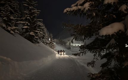 Eine Gruppe von Menschen steht im Schnee und hält Laternen in der Dämmerung. Im Hintergrund sind Berge und eine Schneelandschaft zu sehen.