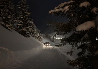 A group of people stands in the snow holding lanterns in the twilight. In the background, mountains and a snowy landscape are visible.