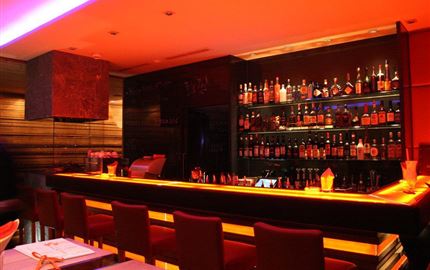 Eine stilvolle Bar mit roten und orangefarbenen Beleuchtungselementen. Regale voller Spirituosen sind im Hintergrund sichtbar.