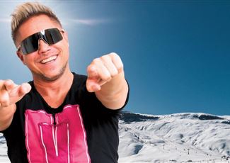 A man with sunglasses and a casual outfit poses in front of a snow-covered landscape. The sky is clear and blue.