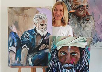 Eine Künstlerin präsentiert ihre Gemälde von Männern mit markanten Bärten und grauen Haaren. Im Hintergrund sind weitere Kunstwerke zu sehen.