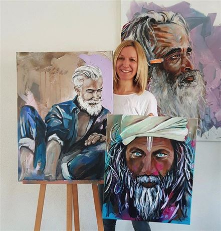 Eine Künstlerin präsentiert ihre Gemälde von Männern mit markanten Bärten und grauen Haaren. Im Hintergrund sind weitere Kunstwerke zu sehen.
