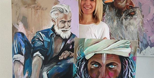 Eine Künstlerin präsentiert ihre Gemälde von Männern mit markanten Bärten und grauen Haaren. Im Hintergrund sind weitere Kunstwerke zu sehen.