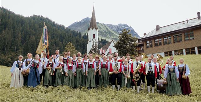 Eine Gruppe von Menschen in traditionellen Trachten steht auf einer Wiese vor einer Kirche und einem Gebäude in den Bergen. Im Hintergrund sind grüne Wälder und die Berglandschaft zu sehen.