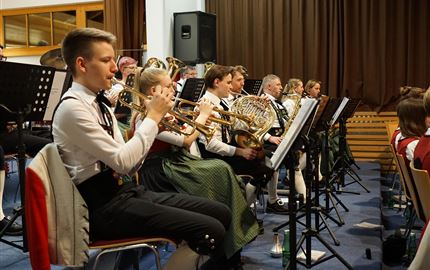 Eine Gruppe von Menschen in traditioneller Tracht steht auf einer Wiese vor einer Kirche und einem Gebäude in den Bergen. Die Umgebung ist grün und ländlich, mit majestätischen Bergen im Hintergrund.