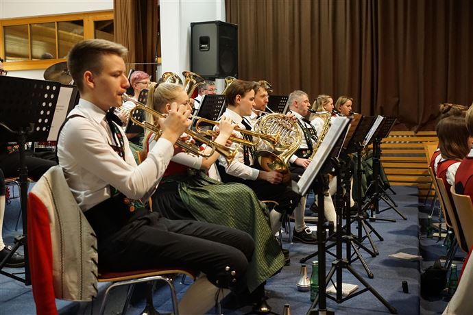 Eine Gruppe von Menschen in traditioneller Tracht steht auf einer Wiese vor einer Kirche und einem Gebäude in den Bergen. Die Umgebung ist grün und ländlich, mit majestätischen Bergen im Hintergrund.