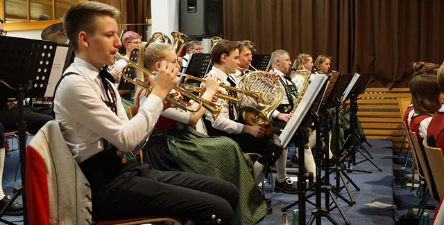 A group of people in traditional costumes stands on a meadow in front of a church and a building in the mountains. The surroundings are green and rural, with majestic mountains in the background.