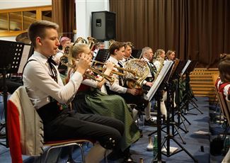 A group of people in traditional costumes stands on a meadow in front of a church and a building in the mountains. The surroundings are green and rural, with majestic mountains in the background.