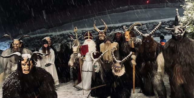 Eine düstere Winternacht mit Menschen in traditionellen Kostümen. Sie tragen Masken und stehen im Schnee, umgeben von einer mystischen Atmosphäre.