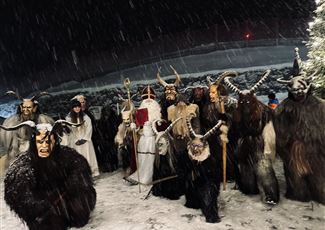 Eine düstere Winternacht mit Menschen in traditionellen Kostümen. Sie tragen Masken und stehen im Schnee, umgeben von einer mystischen Atmosphäre.