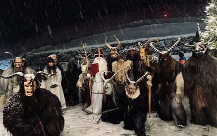 Eine düstere Winternacht mit Menschen in traditionellen Kostümen. Sie tragen Masken und stehen im Schnee, umgeben von einer mystischen Atmosphäre.