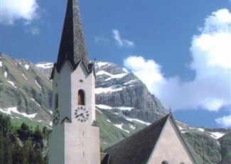 Eine malerische Kirche mit einem hohen Turm, umgeben von Bergen und Wiesen. Der Himmel ist blau mit einigen Wolken.