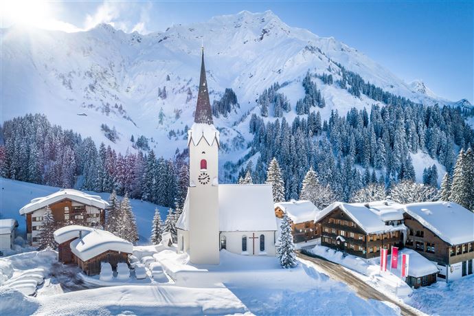 Eine malerische Winterlandschaft mit schneebedeckten Bergen. Im Vordergrund steht eine Kirche mit einem hohen Turm und traditionelle Häuser umgeben von Bäumen.