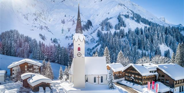 Eine malerische Winterlandschaft mit schneebedeckten Bergen. Im Vordergrund steht eine Kirche mit einem hohen Turm und traditionelle Häuser umgeben von Bäumen.