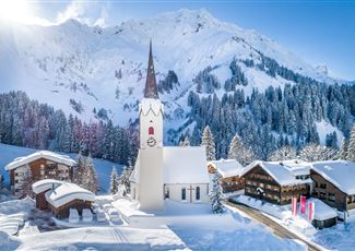 Eine malerische Winterlandschaft mit schneebedeckten Bergen. Im Vordergrund steht eine Kirche mit einem hohen Turm und traditionelle Häuser umgeben von Bäumen.