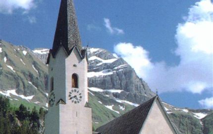 Eine malerische Kirche mit einem hohen Turm, umgeben von Bergen und Wiesen. Der Himmel ist blau mit einigen Wolken.