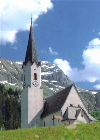 Eine malerische Kirche mit einem hohen Turm, umgeben von Bergen und Wiesen. Der Himmel ist blau mit einigen Wolken.
