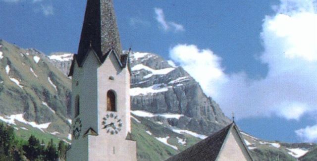 Eine malerische Kirche mit einem hohen Turm, umgeben von Bergen und Wiesen. Der Himmel ist blau mit einigen Wolken.