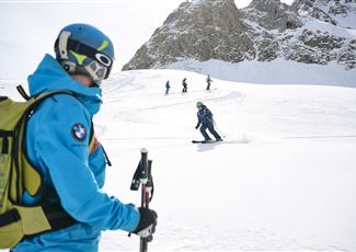 A ski instructor observes students on a snowy slope. In the background, there are other skiers and majestic mountains visible.
