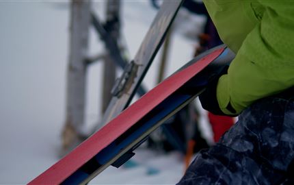 A person is preparing a pair of skis. In the background, a snow-covered landscape and ski equipment are visible.