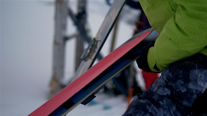 A person is preparing a pair of skis. In the background, a snow-covered landscape and ski equipment are visible.