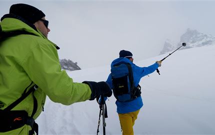 Zwei Männer wandern im Schnee mit Skistöcken. Die Umgebung ist weiß und foggy, umgeben von Bergen.