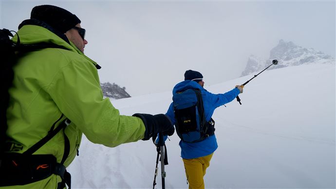 Zwei Männer wandern im Schnee mit Skistöcken. Die Umgebung ist weiß und foggy, umgeben von Bergen.