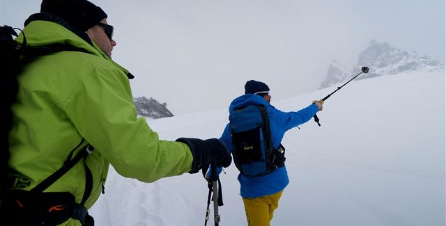 Two men are hiking in the snow with ski poles. The surroundings are white and foggy, surrounded by mountains.
