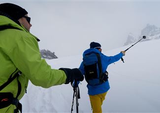 Zwei Männer wandern im Schnee mit Skistöcken. Die Umgebung ist weiß und foggy, umgeben von Bergen.