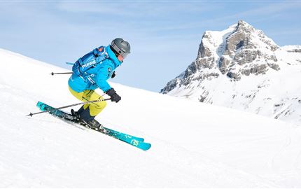 Ein Skifahrer fährt auf einer schneebedeckten Piste. Im Hintergrund sind beeindruckende Berge zu sehen.